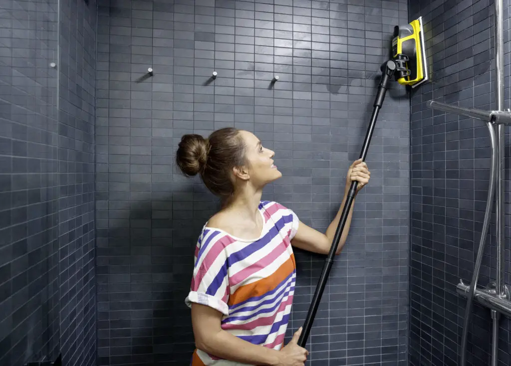 Kobieta czyści czarne płytki w łazience pod prysznicem za pomocą profesjonalnej myjki na lancy