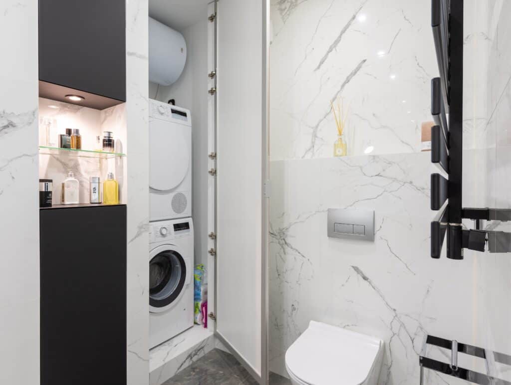 Jasna łazienka z zabudowaną szafą, w której znajduje się suszarka bębnowa i pralka, postawione jedna na drugiej.